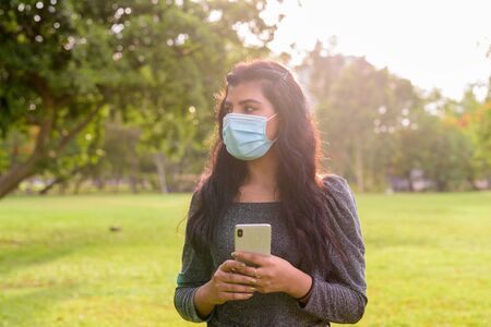 Young Indian woman with mask thinking while using phone at the parkの写真素材