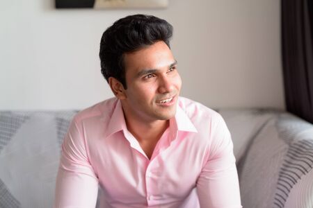 Face of happy young Indian businessman thinking and looking out the window at homeの写真素材