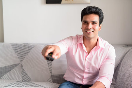 Happy young handsome Indian businessman watching tv at home