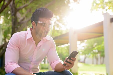 Young handsome Indian businessman with face shield using phone at the parkの写真素材