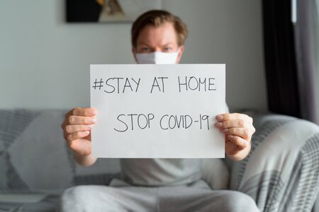 Young man with mask showing Stay At Home sign in the living room while under quarantineの写真素材