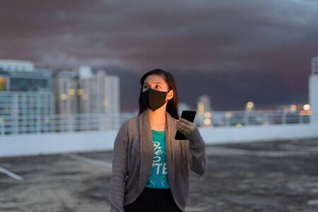 Young Asian woman with mask thinking while using phone during stormy weather outdoorsの写真素材