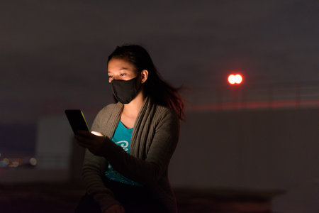 Young Asian woman with mask using phone outdoors during stormy weather at night