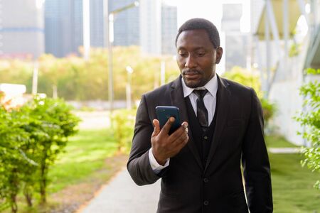 Handsome bearded African businessman in suit using phone at the parkの写真素材