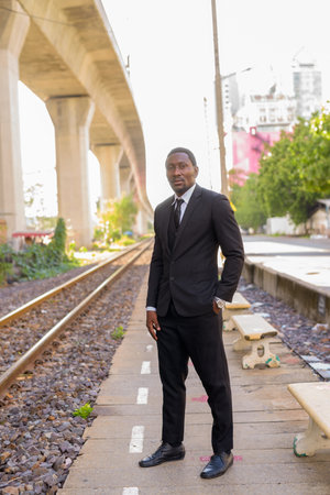 Full body shot of handsome bearded African businessman in suit at the train station outdoorsの写真素材