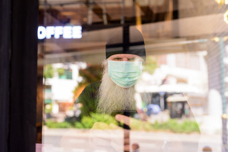 Mature bearded hipster man with mask using phone at the coffee shop through the window