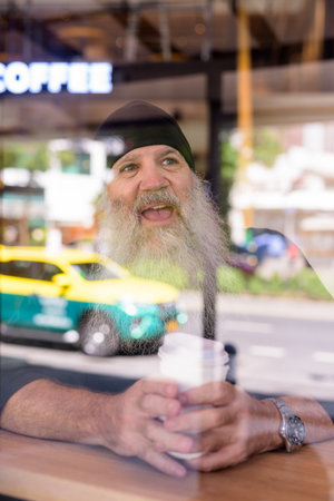 Happy mature handsome bearded hipster man thinking at the coffee shop through the window