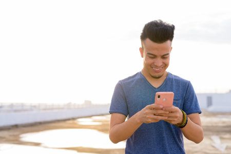 Happy young handsome multi ethnic man using phone at rooftop of the building