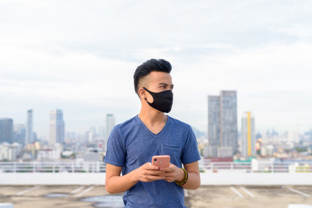 Young multi ethnic man with mask thinking while using phone against view of the cityの写真素材