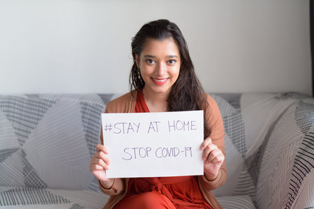 Happy young beautiful Indian woman showing Stay At Home sign while under quarantine due to covid-19...