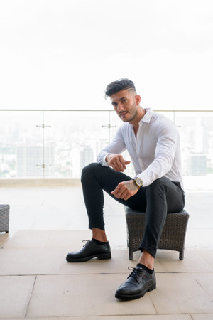 Happy young handsome bearded Persian businessman sitting in the city outdoors