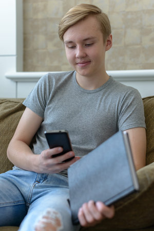 Happy young handsome blond teenage boy holding book while using phone at home