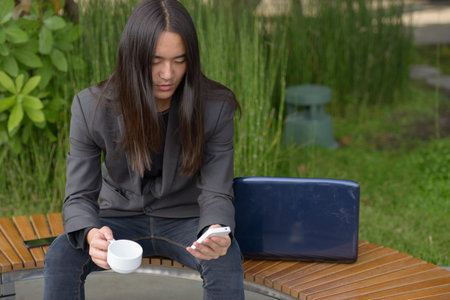 Young Asian businessman with long hair using phone and having coffee at the park