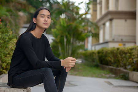 Young Asian man with long hair sitting and smoking at the parkの写真素材