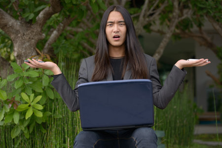 Stressed young Asian businessman with long hair using laptop and two phones while working at the...