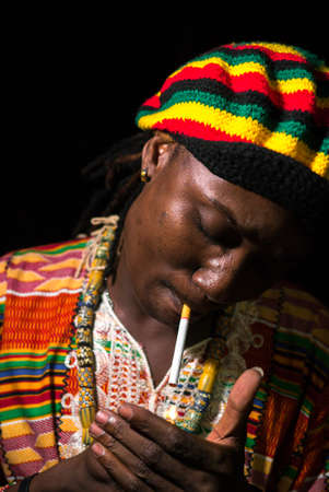 Face of young African Rastafarian man lighting cigarette outdoors at nightの写真素材