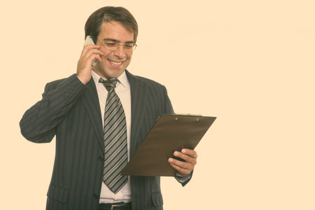 Young happy Persian businessman smiling and talking on mobile phone while reading on clipboard