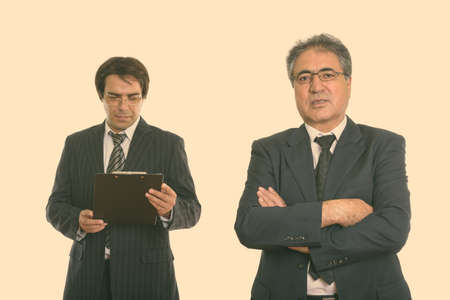 Senior Persian businessman with arms crossed and young Persian businessman reading on clipboardの写真素材