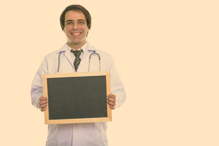 Studio shot of young happy Persian man doctor smiling while holding blank blackboardの写真素材