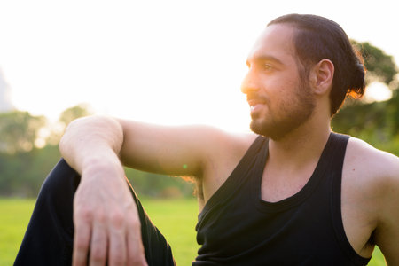 Profile view of young handsome bearded Indian man sitting at the park