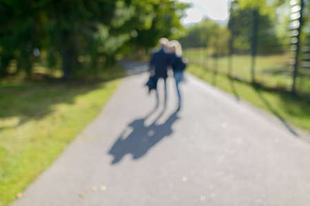 Defocused couple walking in the park along path of green lush trees on sunny dayの写真素材