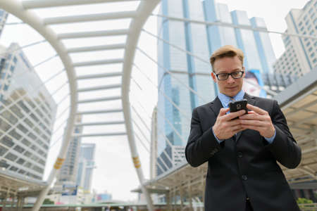 Young handsome blond businessman with eyeglasses using phone in the city outdoorsの写真素材