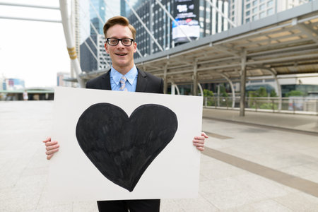 Happy young handsome blond businessman holding heart sign in the city outdoors