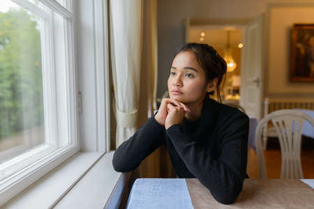 Portrait of young beautiful Asian woman looking out the window at homeの写真素材