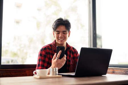 Happy young handsome Asian hipster man using phone and laptop at the coffee shopの写真素材