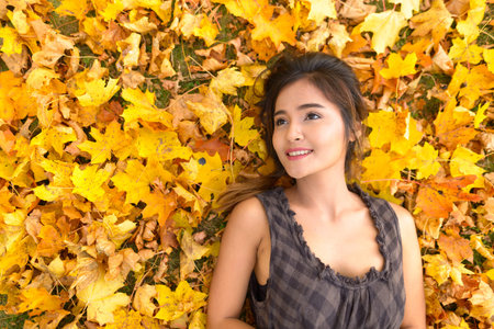 Young happy Asian woman smiling and thinking while lying down on yellow autumn leaves