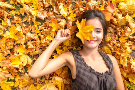 Young happy Asian woman smiling while covering face with leaf lying down on yellow autumn leavesの写真素材