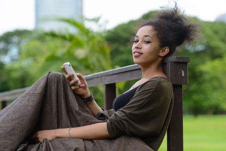 Portrait of happy young beautiful African woman using phone at the parkの写真素材