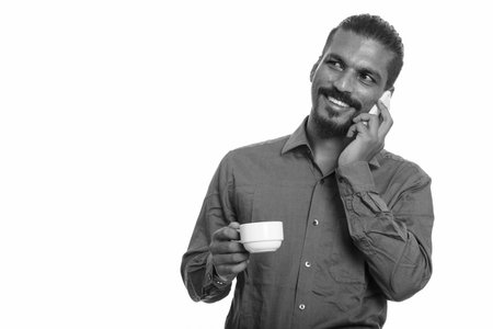 Young happy Indian businessman talking on mobile phone and holding coffee cup while thinking