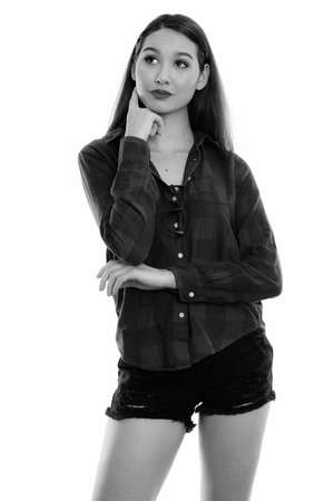 Studio shot of young beautiful Asian woman standing while thinking and looking upの写真素材