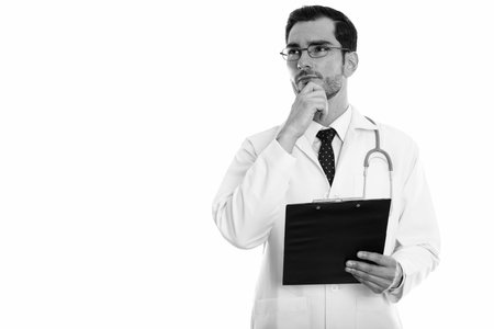 Studio shot of young handsome man doctor thinking while looking up and holding clipboard
