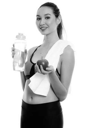 Studio shot of young happy beautiful Asian woman smiling while holding water bottle and apple ready for gymの写真素材