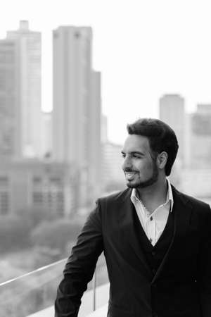 Thoughtful happy Indian businessman smiling while enjoying the view at rooftop against view of the cityの写真素材