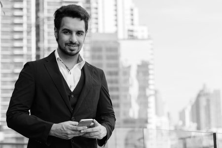 Young happy Indian businessman smiling while holding mobile phone at rooftop against view of the...