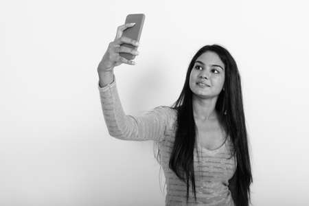Studio shot of young happy Indian woman smiling while taking selfie picture with mobile phone against white backgroundの写真素材