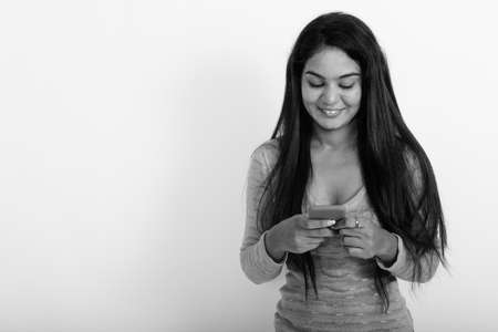 Studio shot of young happy Indian woman smiling while using mobile phone against white backgroundの写真素材