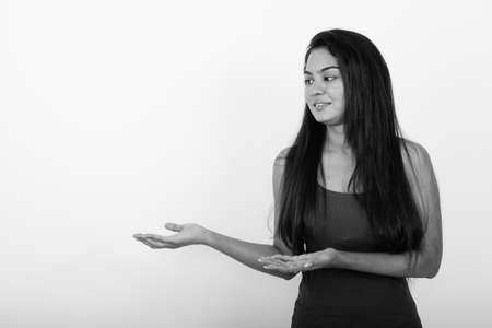 Studio shot of young happy Indian woman smiling while looking to the side and showing something against white backgroundの写真素材