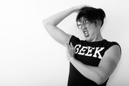 Studio shot of young funny nerd man looking disgusted while smelling armpit and wearing eyeglasses