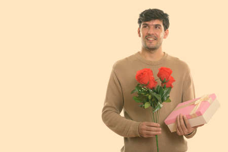 Thoughtful young happy Persian man smiling while holding red roses and gift box ready for Valentines dayの写真素材