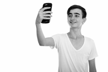 Studio shot of young happy Persian teenage boy smiling while taking selfie picture with mobile phoneの写真素材