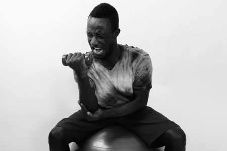Young African fitness man sitting on exercise ball while doing bicep curl with light weight dumbbell