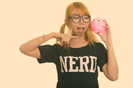 Young happy Asian nerd woman smiling and holding piggy bank while pointing fingerの写真素材