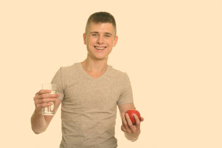 Young happy Caucasian man smiling while holding glass of water and red appleの写真素材