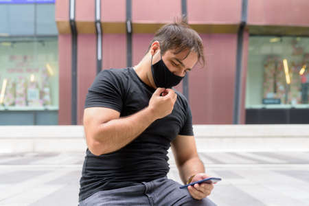 Portrait of Persian man with mask using phone and sitting in the city outdoorsの写真素材