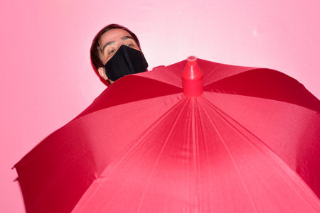 Persian businessman with mask holding umbrella against pink wallの写真素材