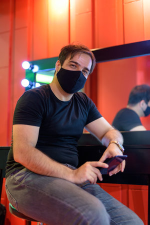 Persian man with mask holding phone while sitting against mirror in the makeup room